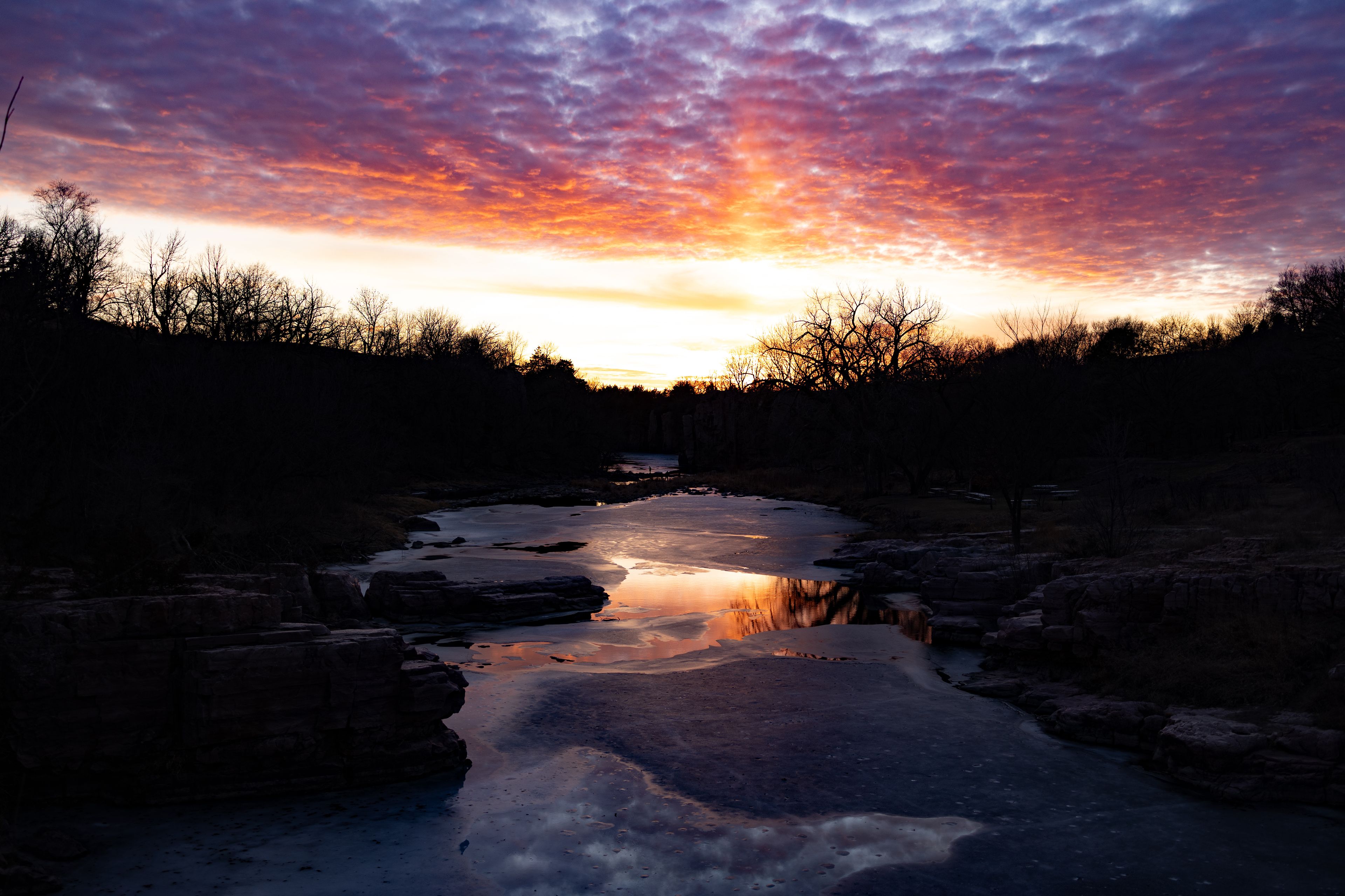 Tramonto su un fiume con sponda rocciosa e alberi in controluce.