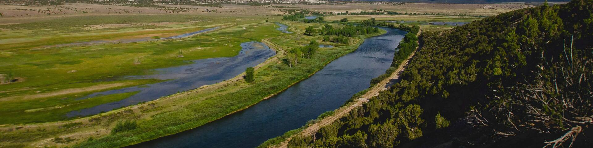 A river winding through a valley with green fields and distant mountains under a partly cloudy sky.