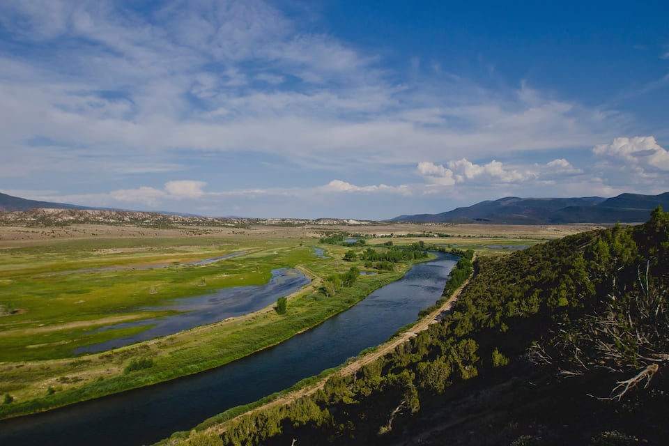 A river winding through a valley with green fields and distant mountains under a partly cloudy sky.