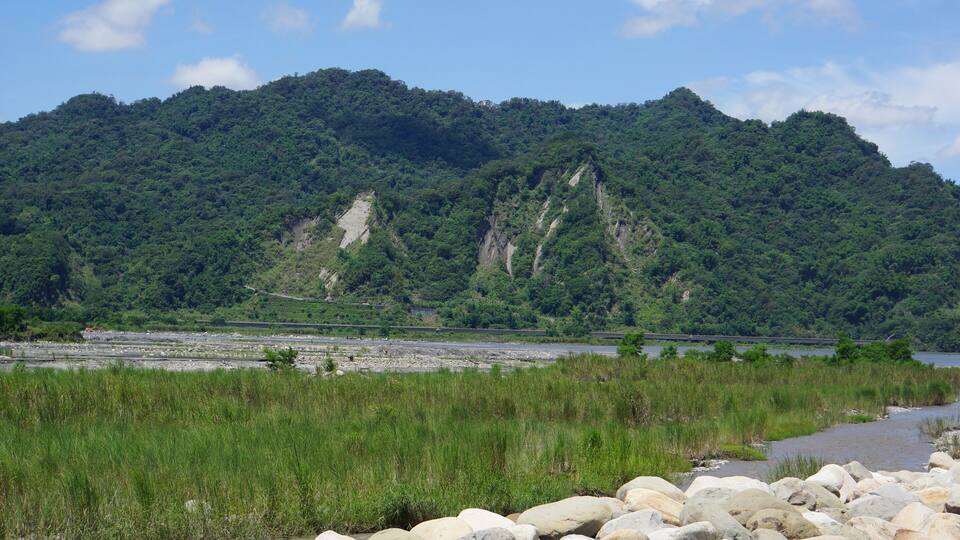 A river with rocks in the foreground, a forested hill in the background, and a clear sky.