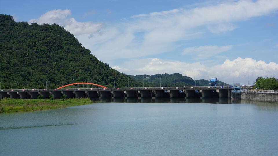 A bridge over a calm river with a forested hill in the background.