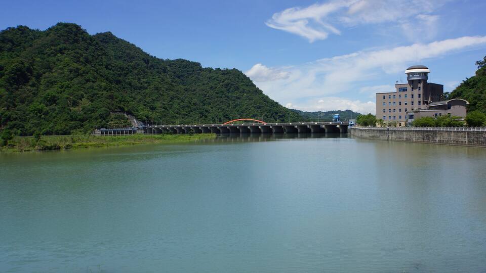 A bridge over a calm river, surrounded by lush green hills and a large building with a tower.