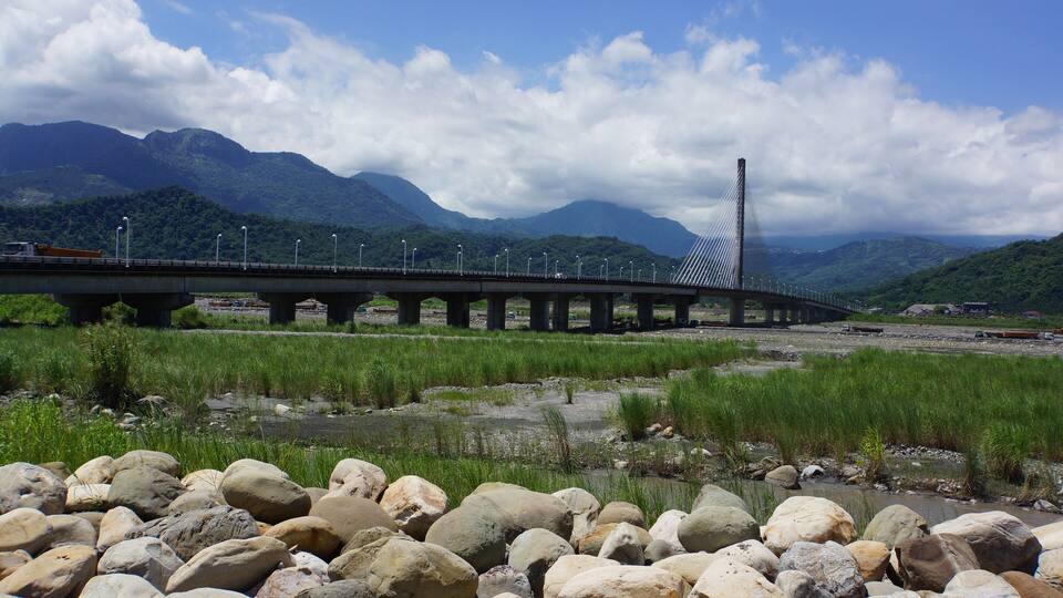 A bridge with a modern design, a river with rocks, and mountains in the background.