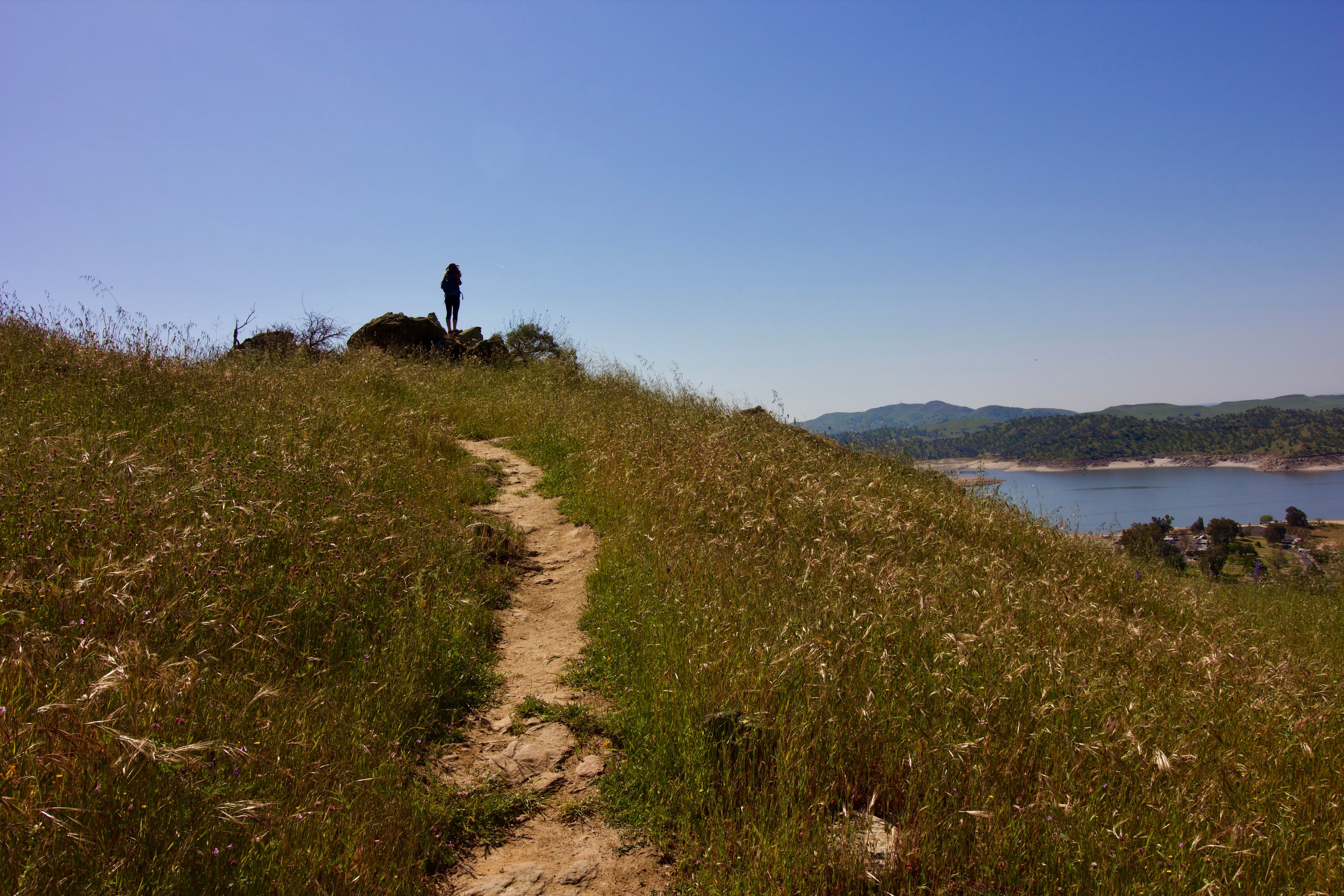 A dirt path leading to a rocky outcrop with a person standing on it, overlooking a body of water and distant hills.