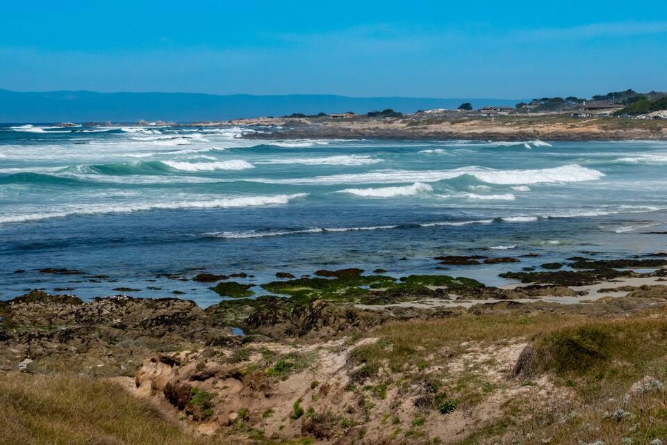 Een kustlandschap met golven die op rotsachtige oevers breken, een zandstrand en heuvels in de verte onder een heldere lucht.