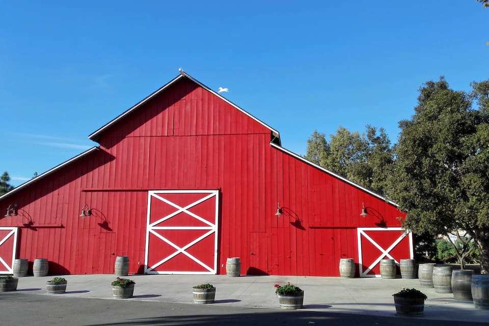A red barn with white cross doors and windows, surrounded by potted plants and barrels.