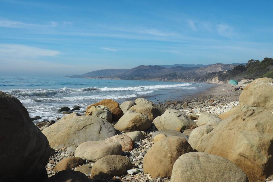 Une plage rocheuse parsemée de gros rochers, des vagues qui s’écrasent sur le rivage et, au loin, des collines sous un ciel dégagé.