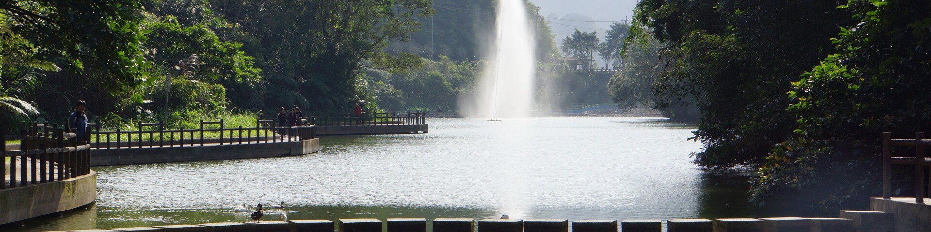 Un lago sereno con una fuente central, rodeado de exuberante vegetación y colinas.