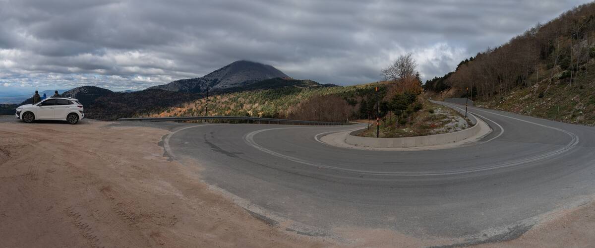 A winding road with a white car parked on the left, surrounded by a mountainous landscape and autumn-colored trees.