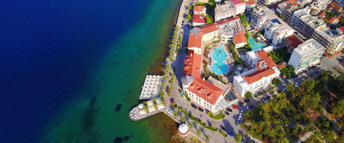 An aerial view of a coastal town with a clear blue sea, a central swimming pool surrounded by buildings, and a small pier.