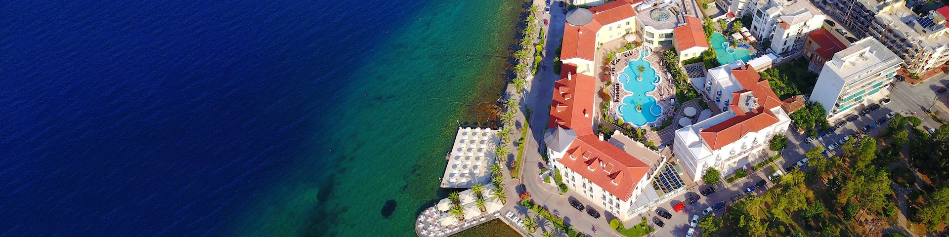 An aerial view of a coastal town with a clear blue sea, a central swimming pool surrounded by buildings, and a small pier.