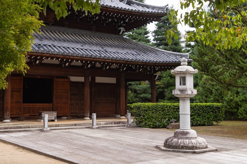 A traditional Japanese building with a tiled roof and wooden structure, surrounded by greenery and a stone lantern.