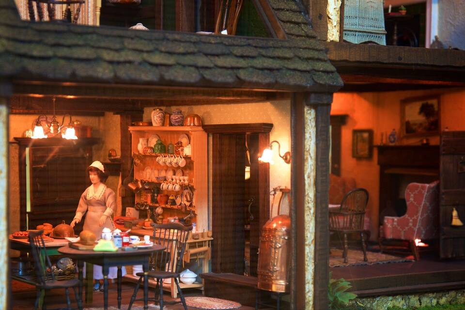 A model of a rustic shop with a woman standing by a table, shelves with various items, and a large copper pot.