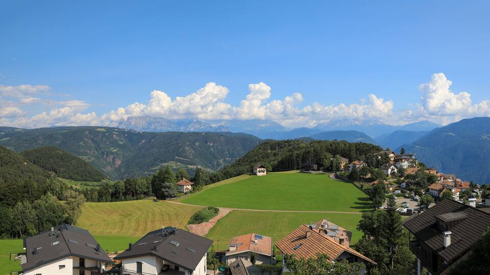 A village nestled in a valley with green fields, houses, and mountains in the background.