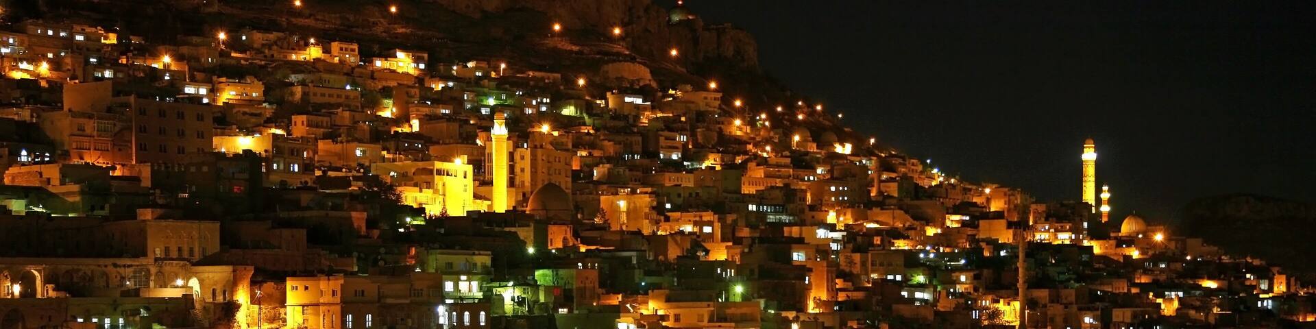 A densely built hillside town at night with illuminated buildings and a prominent minaret.