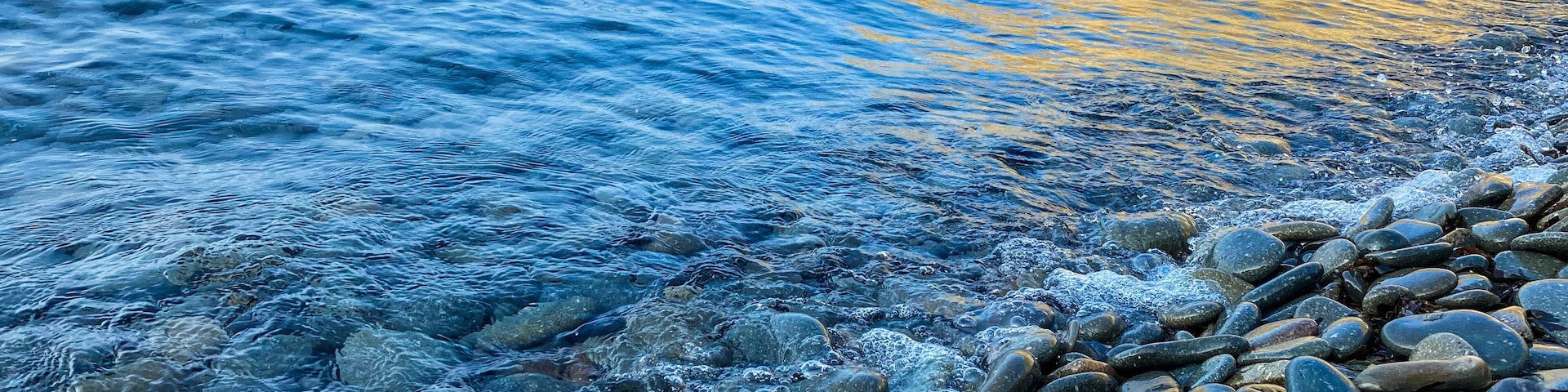 A rocky shoreline with clear water and a golden reflection on the water's surface.