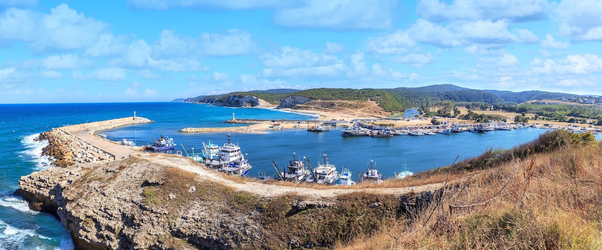 A coastal harbor with boats, rocky cliffs, and a distant mountain range.