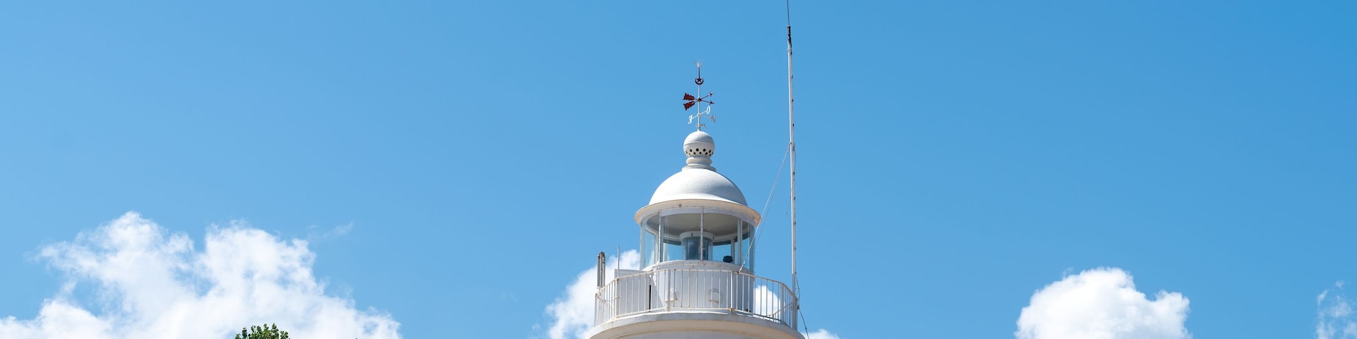Un faro blanco con una veleta en la parte superior, frente a un cielo azul despejado con algunas nubes.