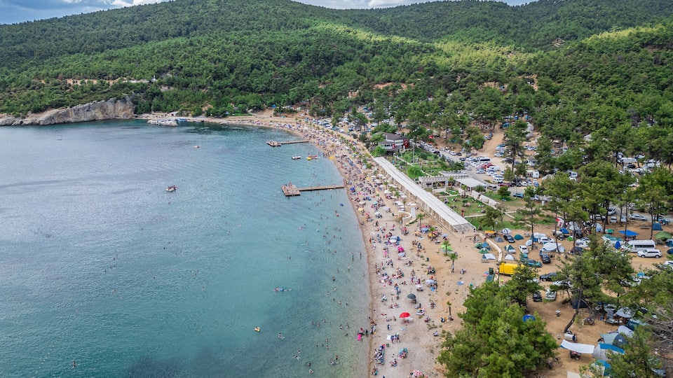 A coastal beach with clear turquoise waters, a sandy shore, and a forested hill in the background.