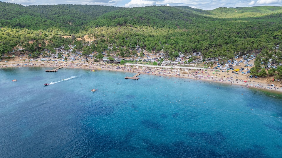 A coastal beach with clear blue waters, a pier, and a mountainous backdrop.