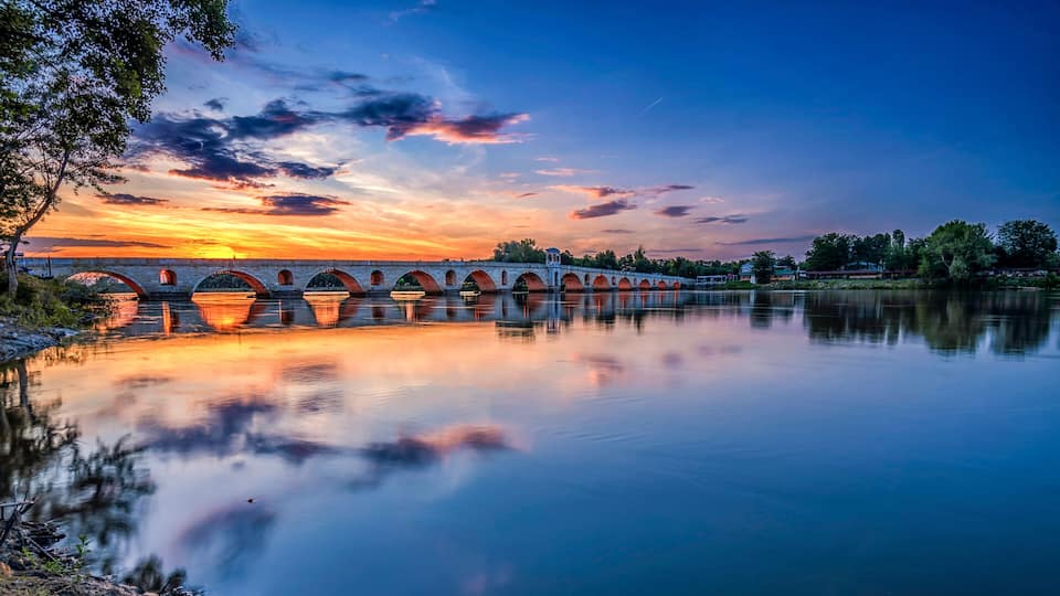 A stone bridge with multiple arches over a calm river, reflecting the sunset sky.
