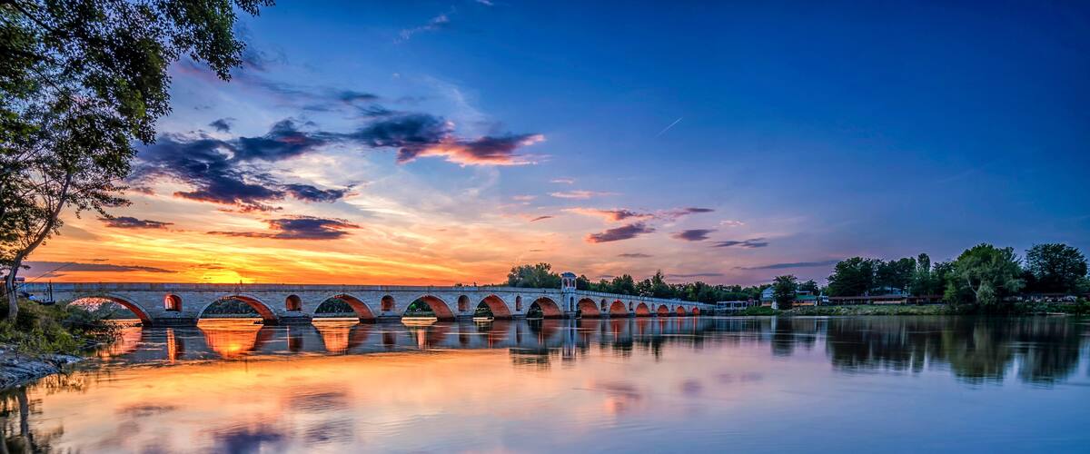A stone bridge with multiple arches over a calm river, reflecting the sunset sky.