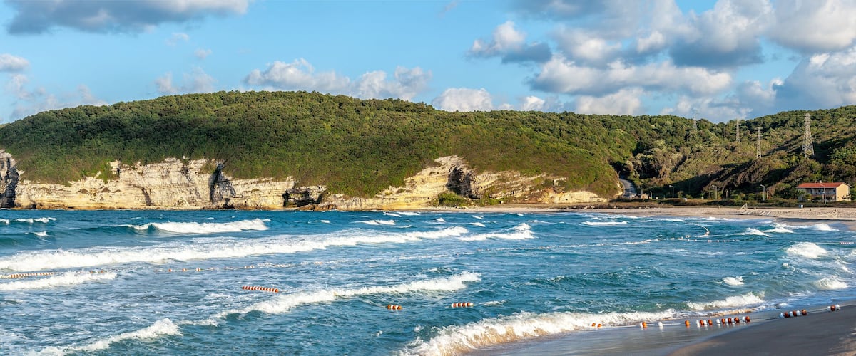 A beach with waves, a rocky cliff, and a forested hill.