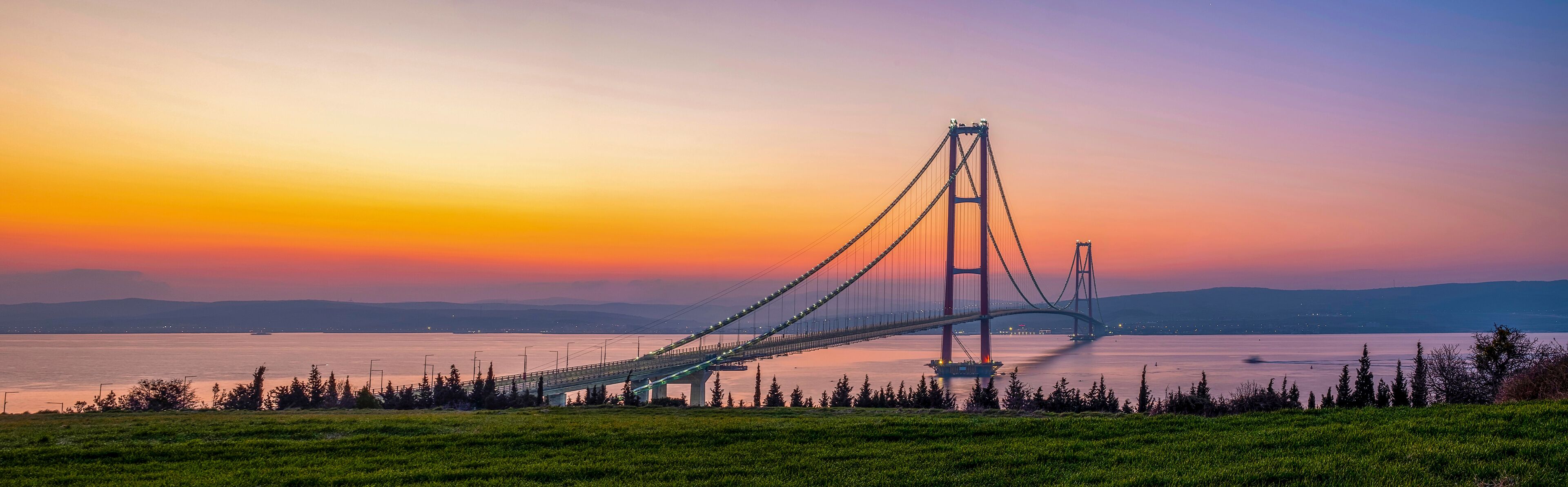 A suspension bridge at sunset with a clear sky and calm water.