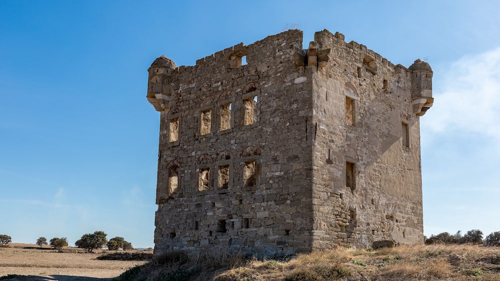 A large, weathered stone tower with multiple windows and a flat roof, surrounded by dry grass and sparse trees.