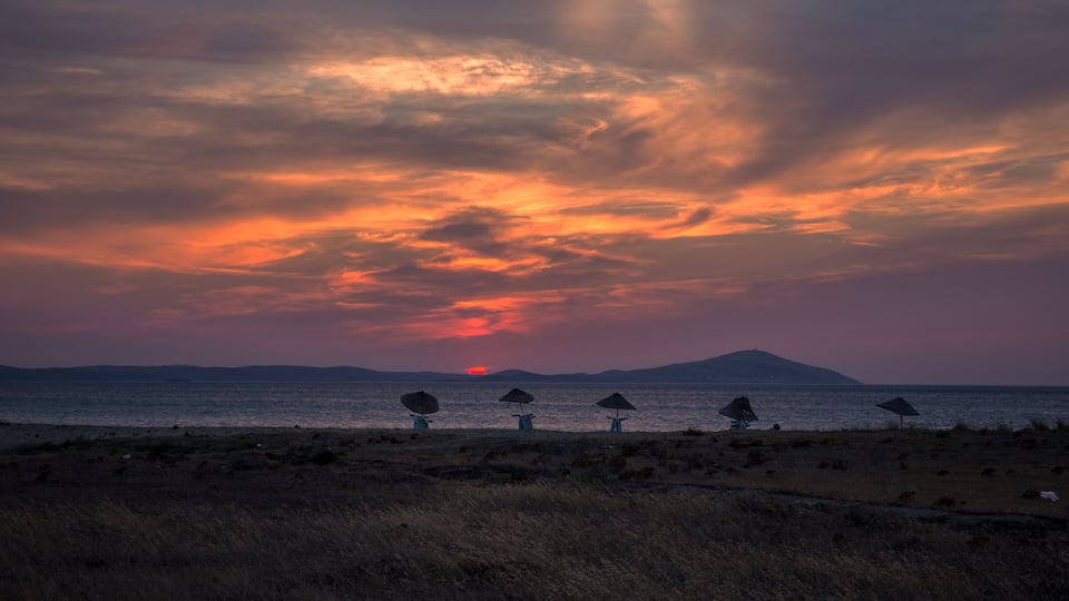 A sunset over a body of water with a distant mountain and several beach umbrellas.