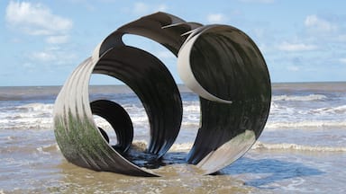 Une grande sculpture métallique composée de trois sections incurvées, située sur une plage avec des vagues en arrière-plan.