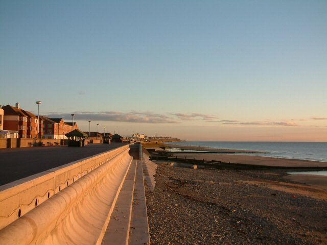 A coastal promenade with buildings on the left, a sandy beach, and the ocean in the background.