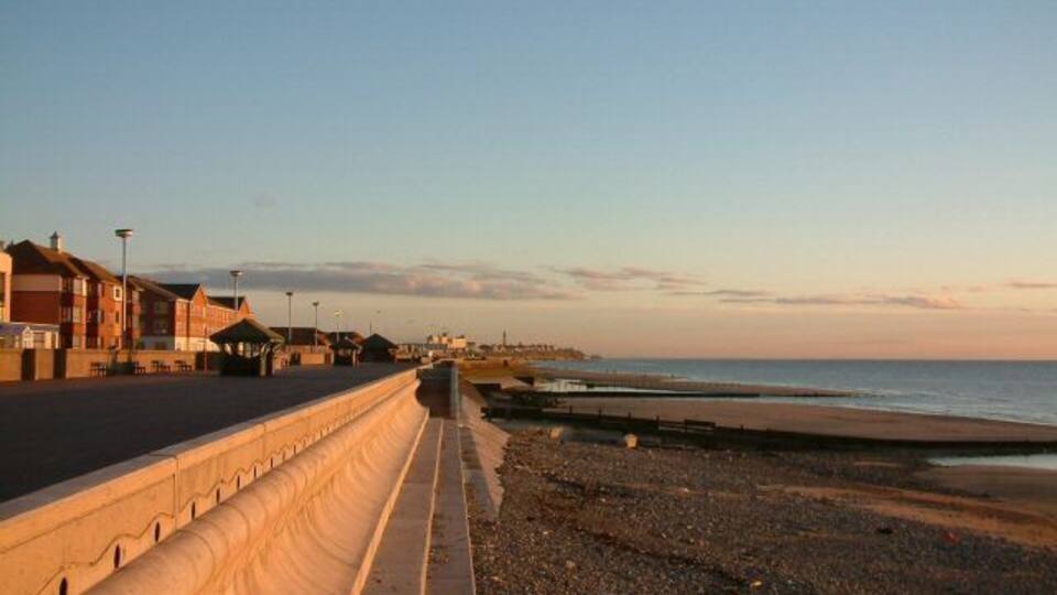 A coastal promenade with buildings on the left, a sandy beach, and the ocean in the background.
