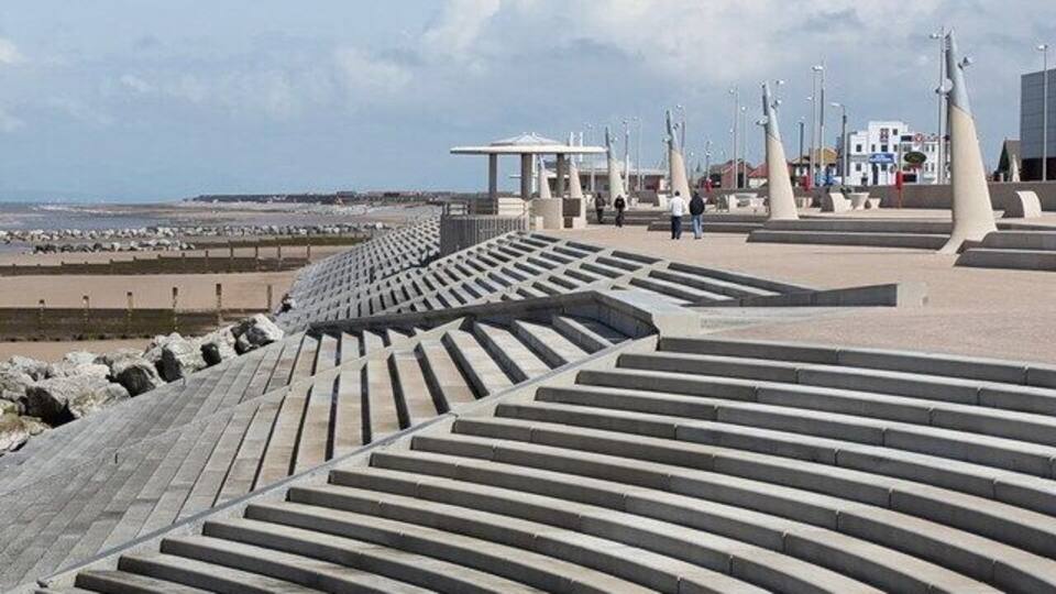 A coastal promenade with steps leading to the beach, a white gazebo, and modern sculptures.