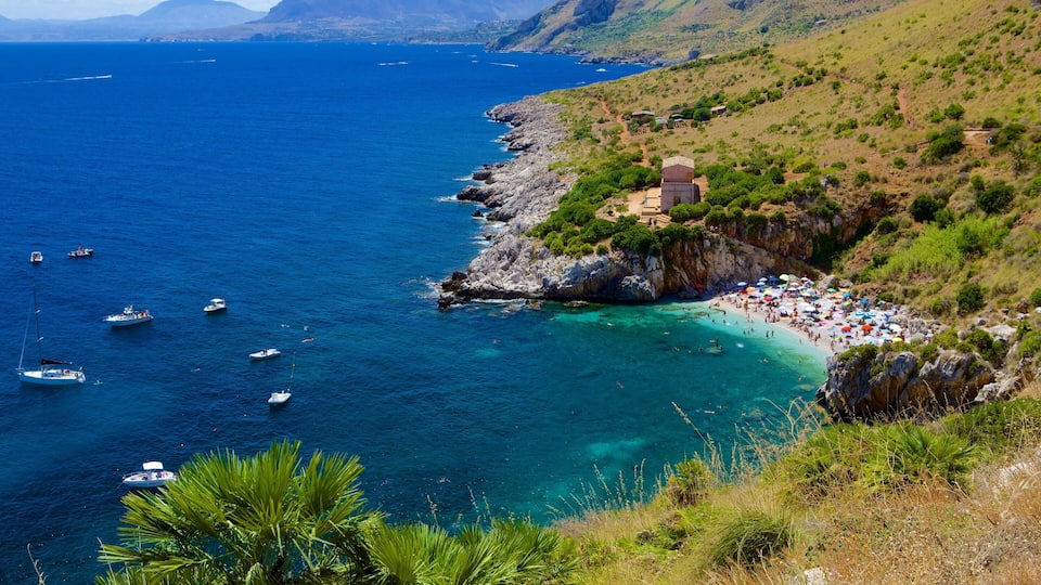 A coastal landscape with a rocky shoreline, clear blue waters, and a beach populated with umbrellas and people.