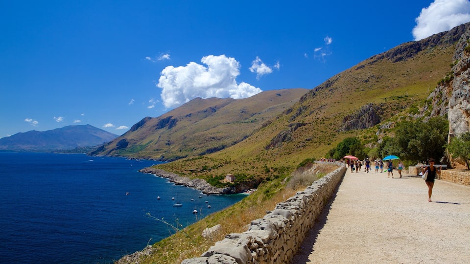 A coastal path with people walking, a stone wall, and boats in the water.