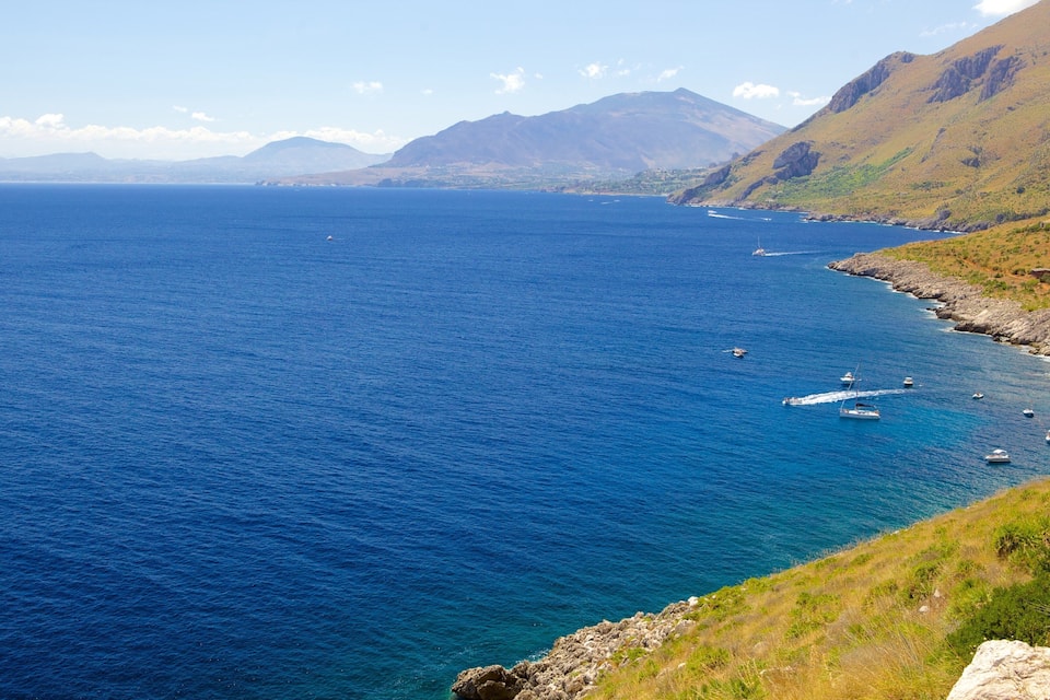 A coastal landscape with a clear blue sea, rocky coastline, and mountains in the background.