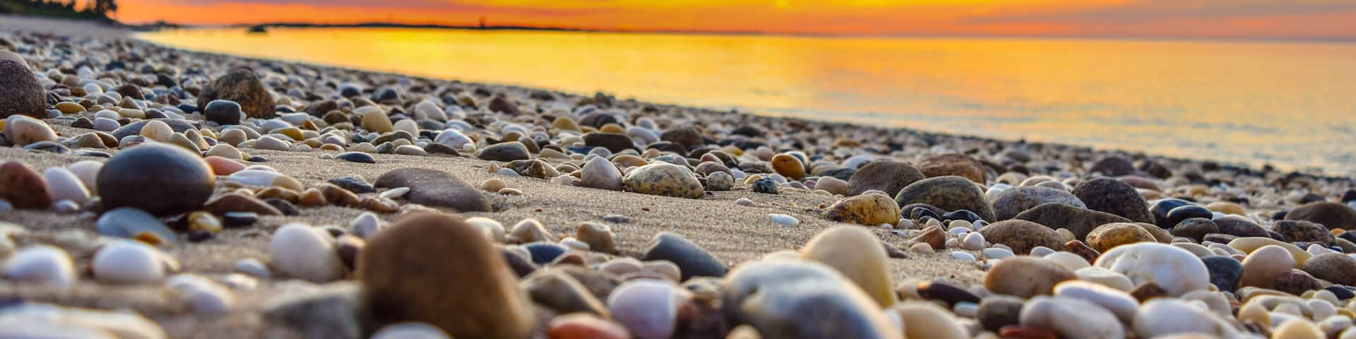 A pebbled beach at sunset with a calm sea and a clear sky.