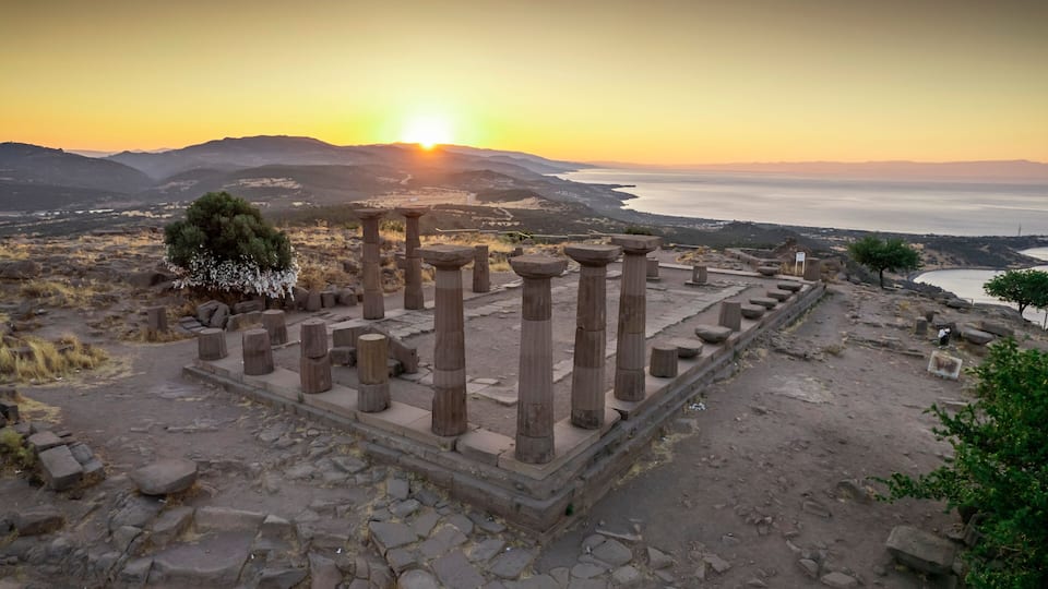 Ancient ruins with columns, a tree, and a sunset over a coastal landscape.