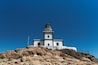 Un phare blanc coiffé d’une partie verte, situé sur une colline rocheuse, sous un ciel bleu dégagé.