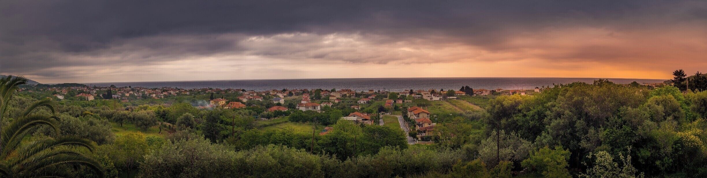 A coastal town with houses and dense greenery, under a dramatic sky.