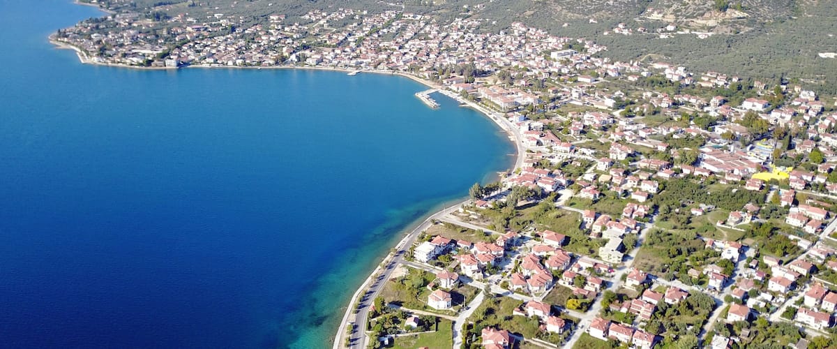 An aerial view of a coastal town with a clear blue sea, surrounded by mountains and residential areas.