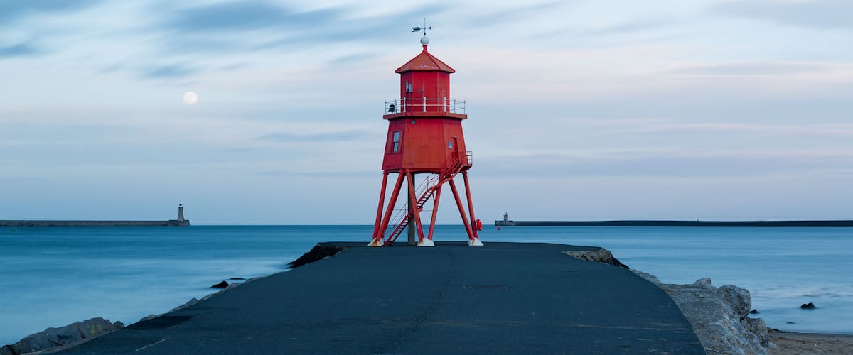 A red lighthouse on a pier with calm water and a distant ship.