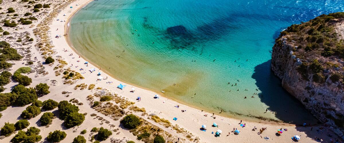 A coastal landscape with a curved bay, clear turquoise waters, sandy beaches, and rocky cliffs.