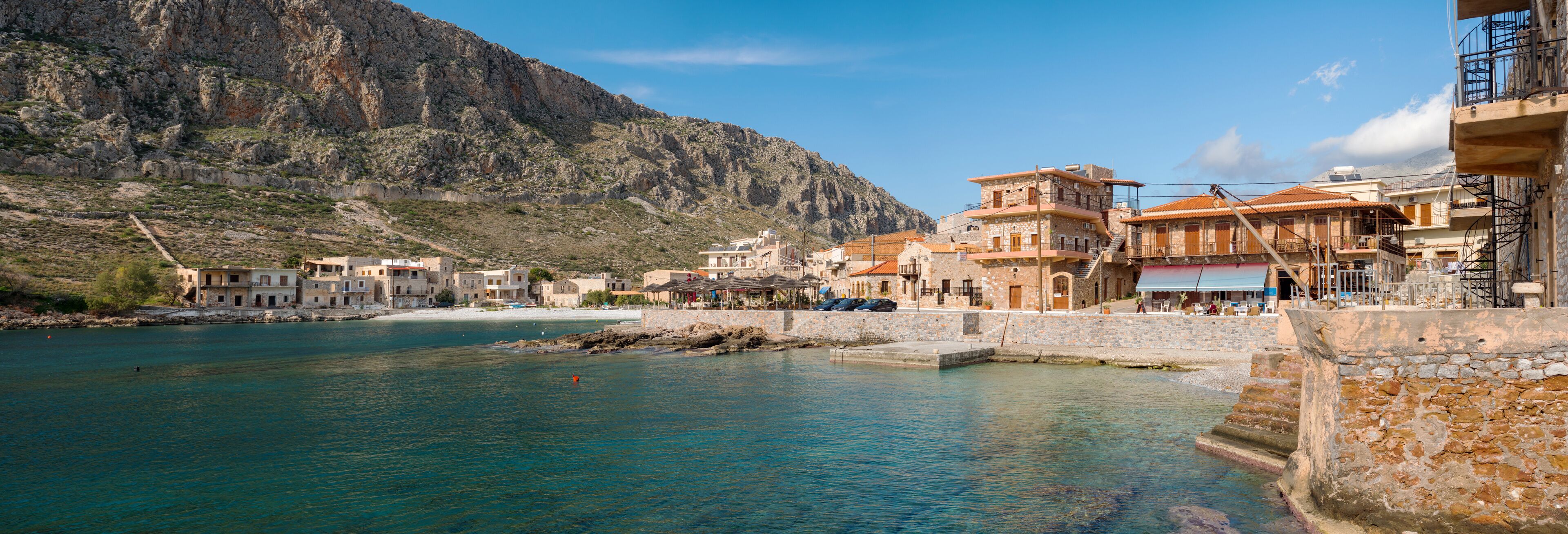 A coastal town with stone buildings, a clear blue sea, and a mountainous backdrop.