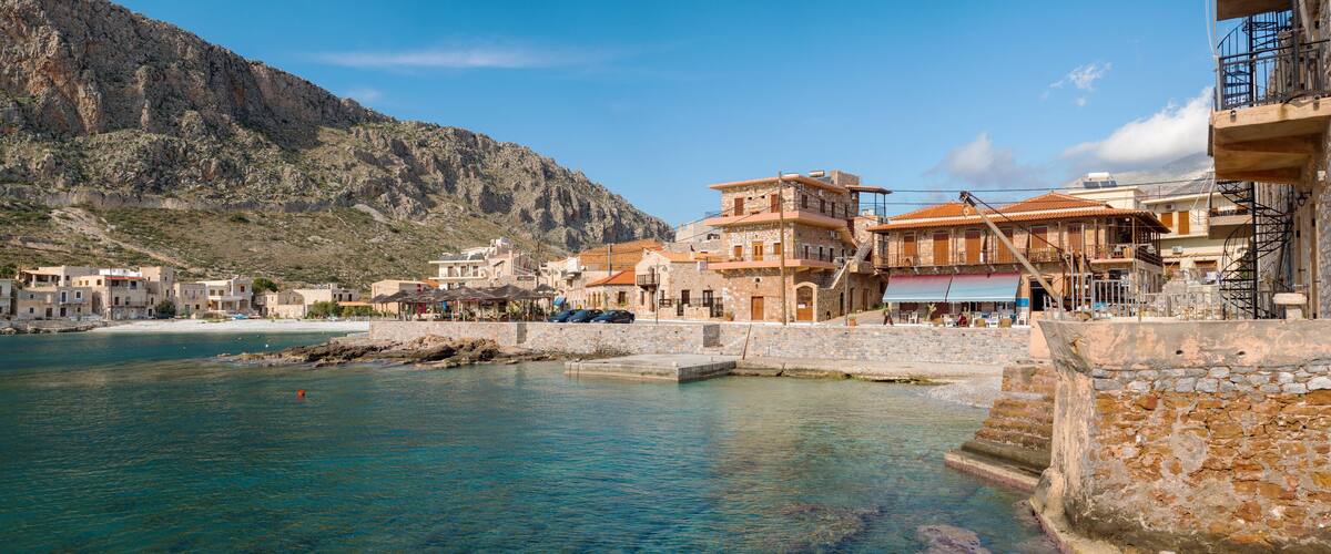 A coastal town with stone buildings, a clear blue sea, and a mountainous backdrop.
