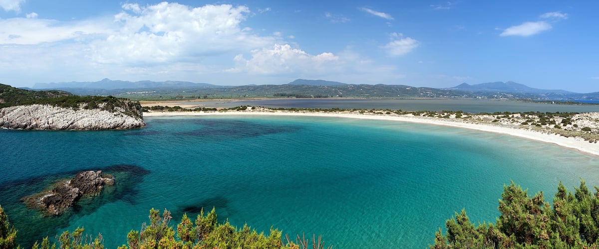 A coastal landscape with clear turquoise waters, a sandy beach, rocky outcrops, and distant mountains under a partly cloudy sky.