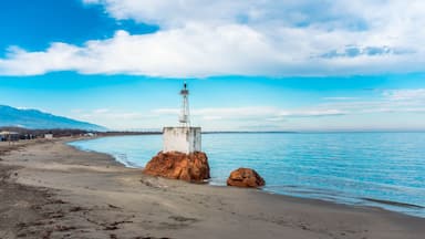 A lighthouse on a rocky outcrop by a beach with a clear sky and mountains in the background.