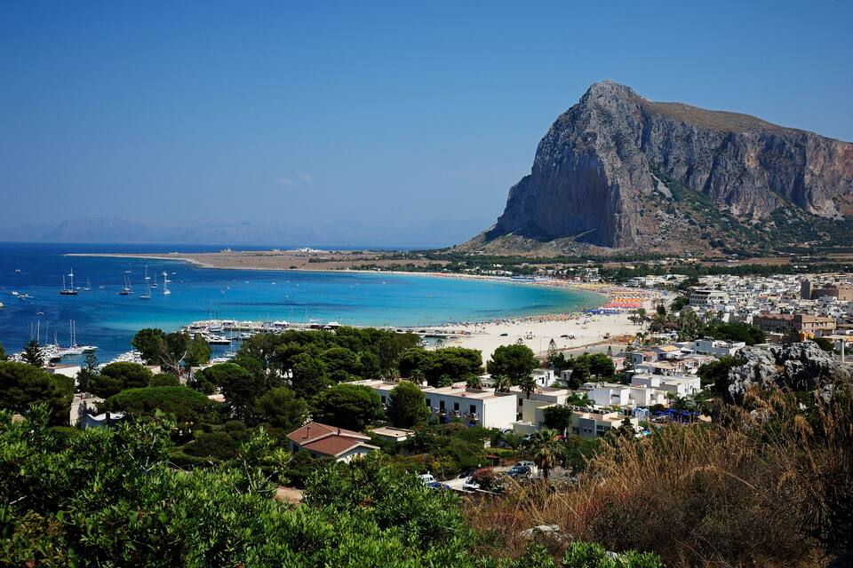 A coastal town with a prominent mountain, clear blue waters, and a sandy beach.