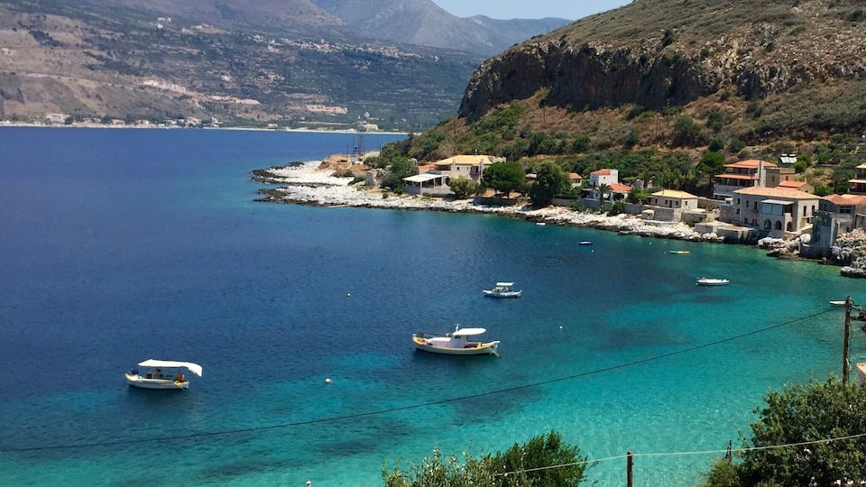 A coastal town with boats on clear turquoise waters, surrounded by mountains and houses.